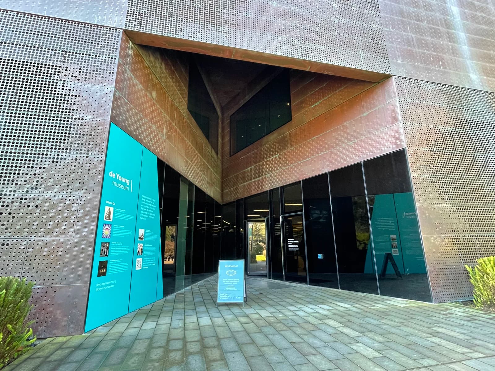 Exterior entrance of the de Young Museum in San Francisco's Golden Gate Park, showing the distinctive copper-clad Herzog & de Meuron building facade