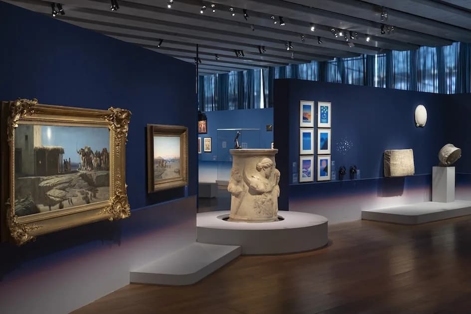 Exhibition view at the MuCEM showing historical paintings, a classical relief, and contemporary works on deep blue walls