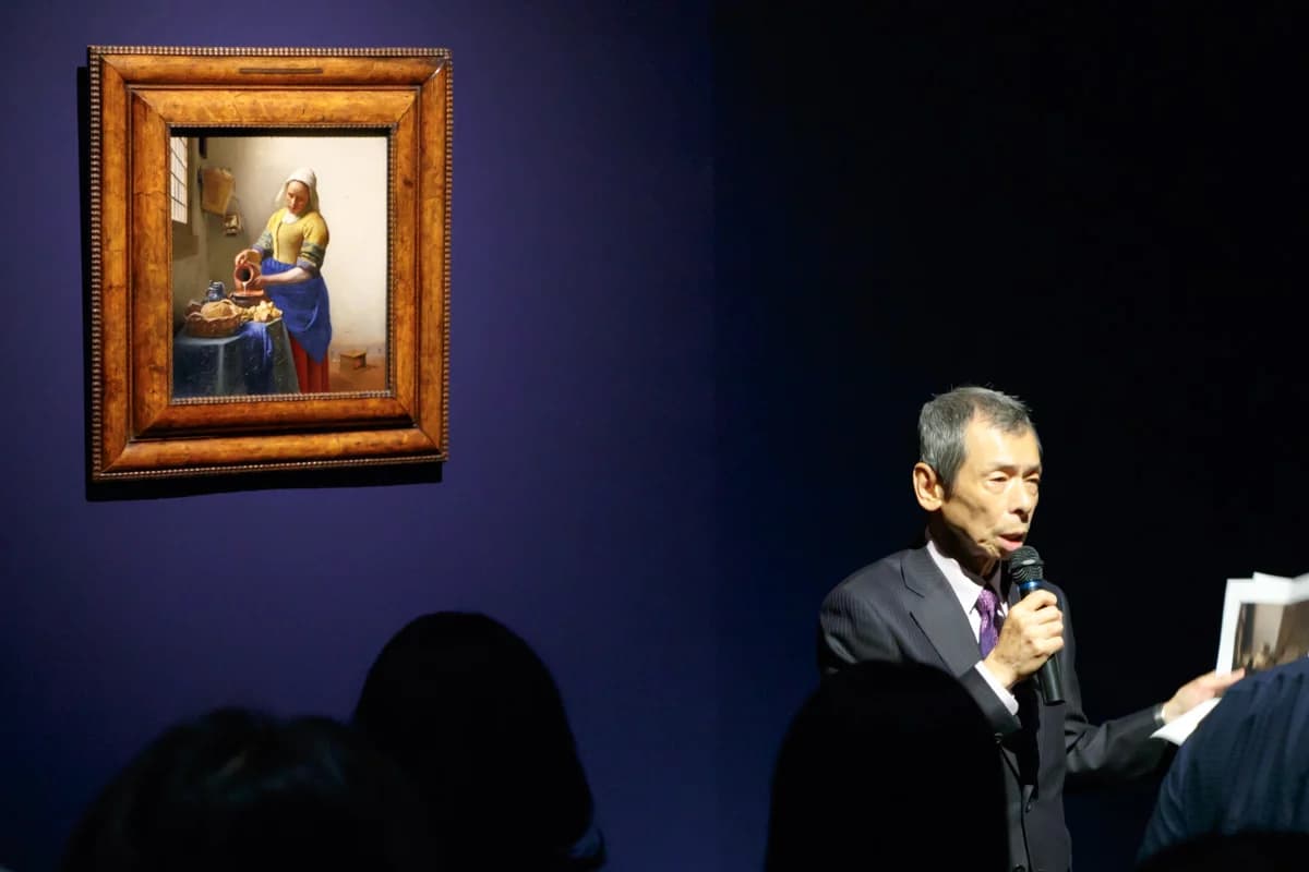 The Milkmaid by Vermeer hanging on a deep-blue wall while a man speaks into a microphone at the press preview