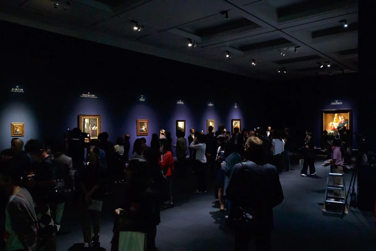 Wide view of the darkened final room with several small Vermeer paintings on a purple wall and a crowd of visitors