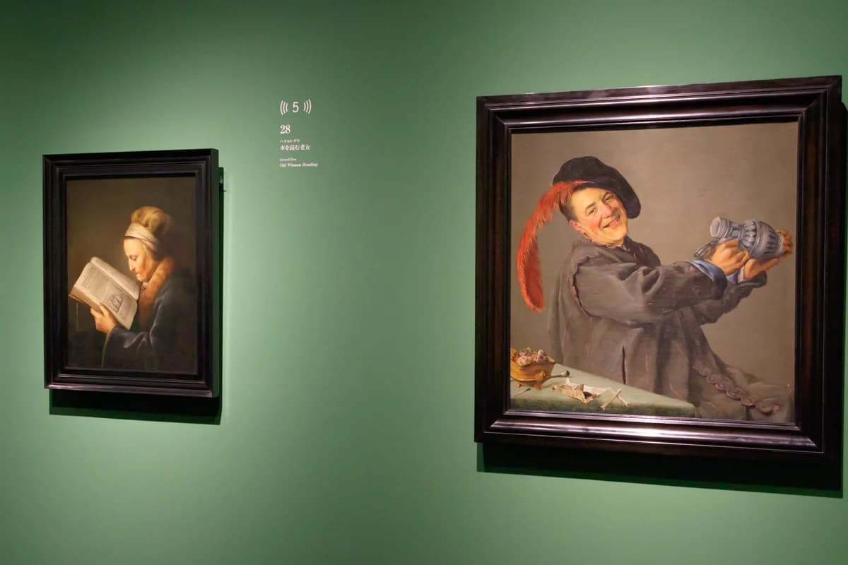 A green gallery wall with Gerard Dou's Old Woman Reading at left and a laughing genre figure at right, with an audio-guide number between them