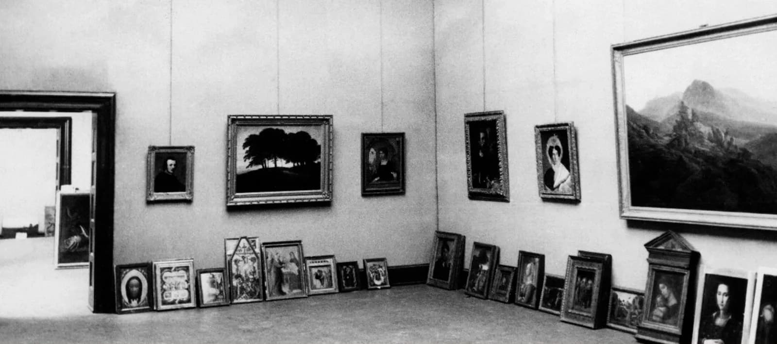 Black-and-white photograph of a gallery room at the Wiesbaden Central Collecting Point in 1945, its high walls hung with framed Old Master paintings and dozens more canvases propped in rows along the floor.