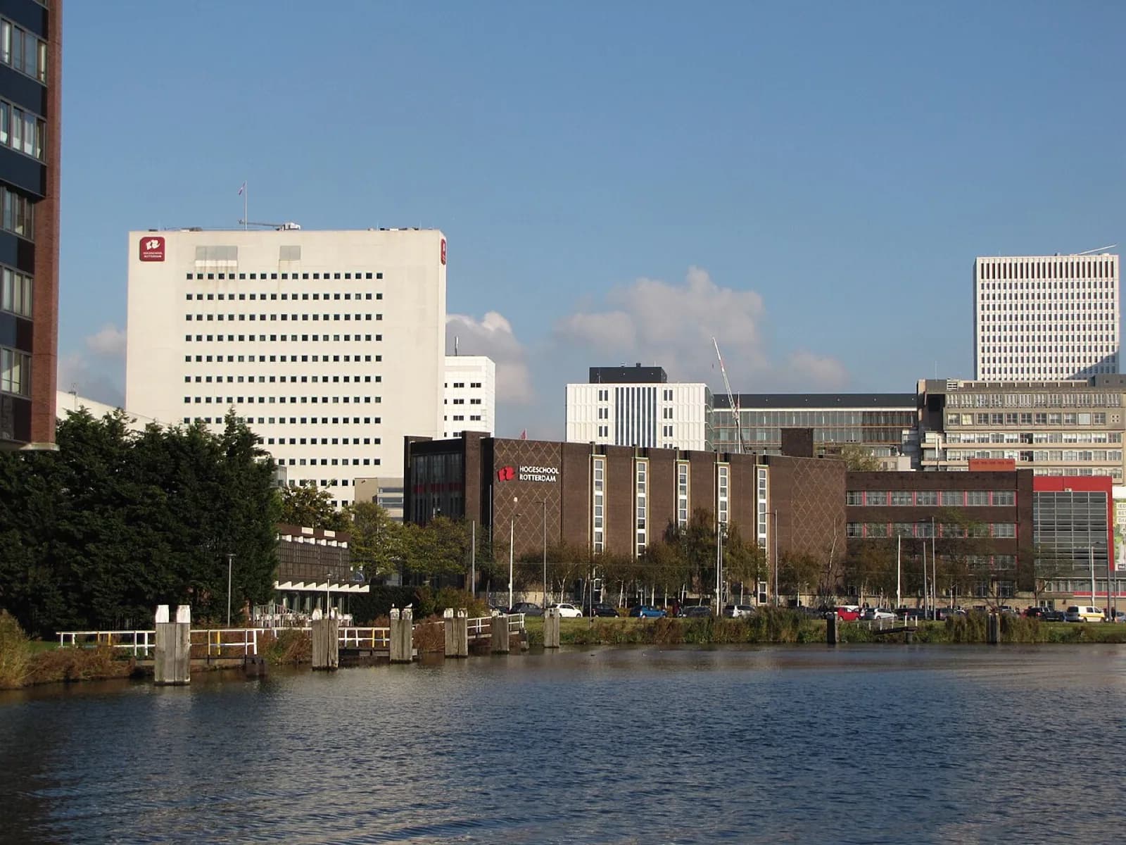 View across the water of the Hogeschool Rotterdam campus buildings including the former Academie van Beeldende Kunsten structures, featuring a prominent white multi-story building with a red logo, adjacent brown brick building, trees, and canal in the foreground under a clear blue sky in Rotterdam, Netherlands.