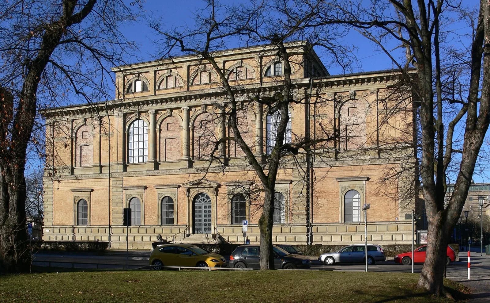 The grand historic facade of the Alte Pinakothek in Munich, a neoclassical building in yellow stone with arched windows and decorative friezes, viewed across a lawn with bare trees and parked cars under a clear blue sky.