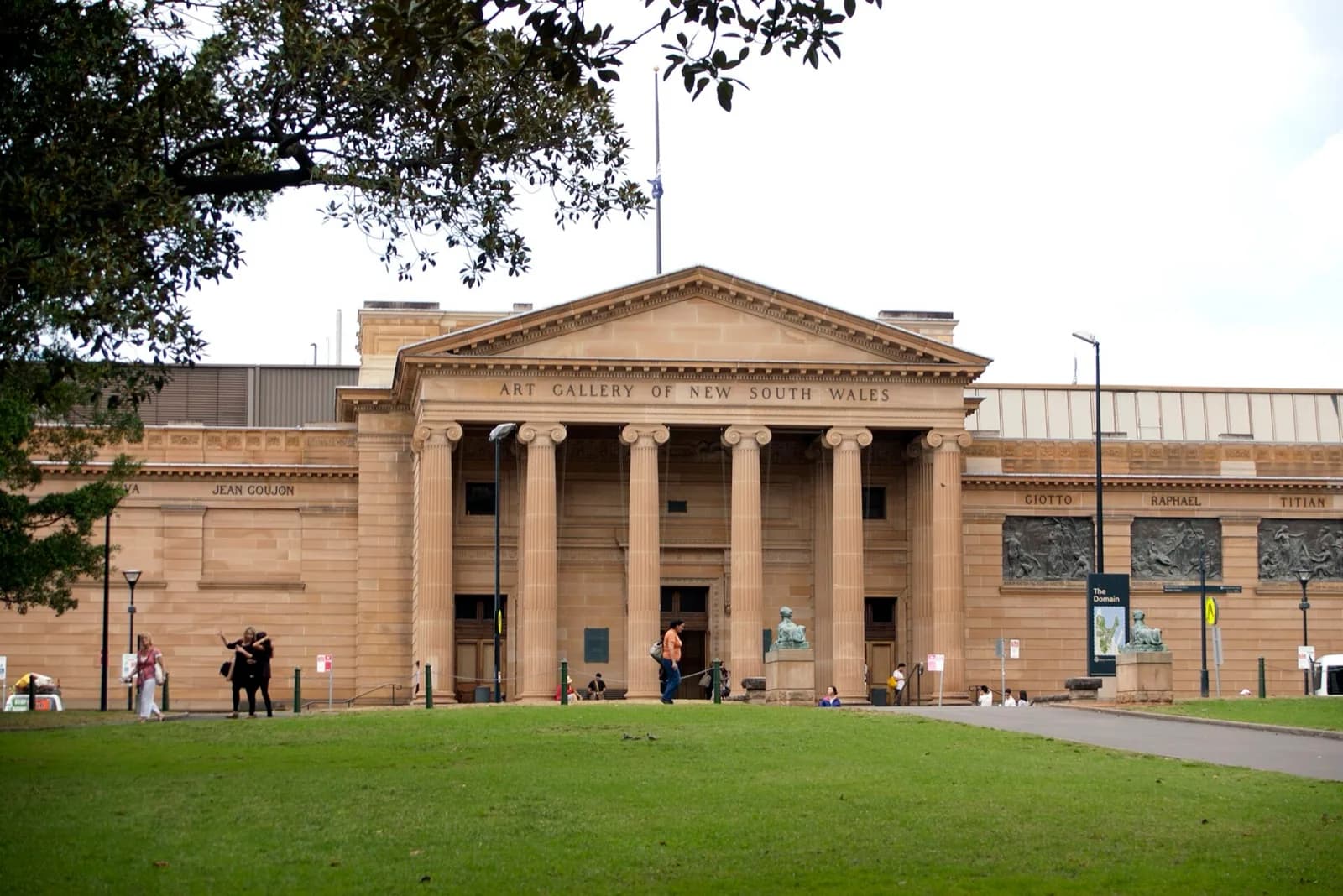 The neoclassical sandstone facade of the Art Gallery of New South Wales in Sydney, featuring its grand columned portico with pediment inscription, sculptural reliefs, and surrounding lawns and trees under a bright sky.