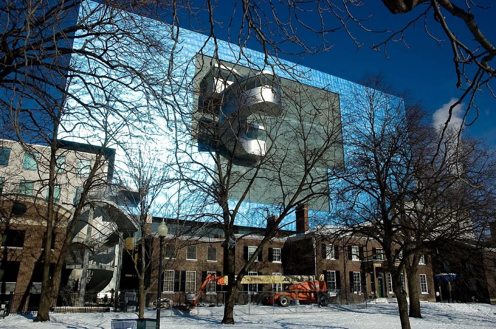 The Art Gallery of Ontario in Toronto on a winter day, showing the historic brick Grange building with the dramatic modern blue glass and stainless steel addition by Frank Gehry rising behind it, viewed across snow-covered Grange Park with bare trees in the foreground under a clear blue sky.