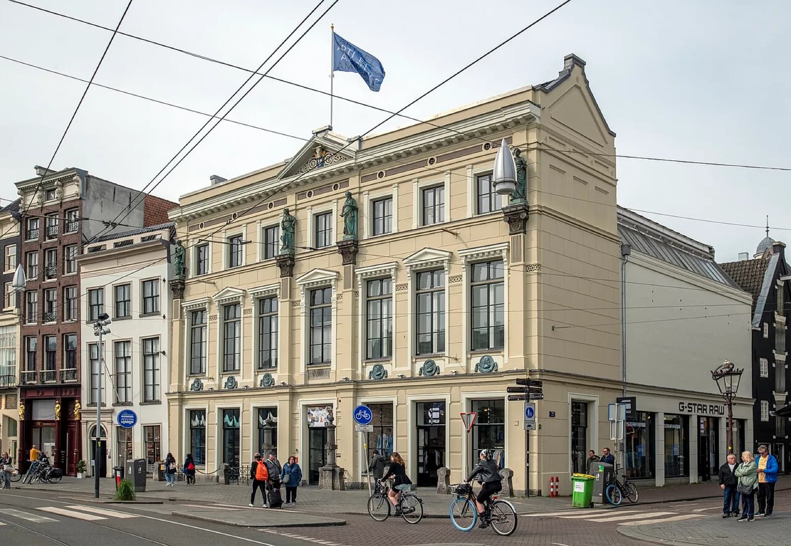 The restored neoclassical cream-colored facade of the Arti et Amicitiae artists' society building on the Rokin in Amsterdam, Netherlands, with ornate statues, decorative reliefs, a blue flag flying, large windows, and pedestrians and cyclists on the street in front.