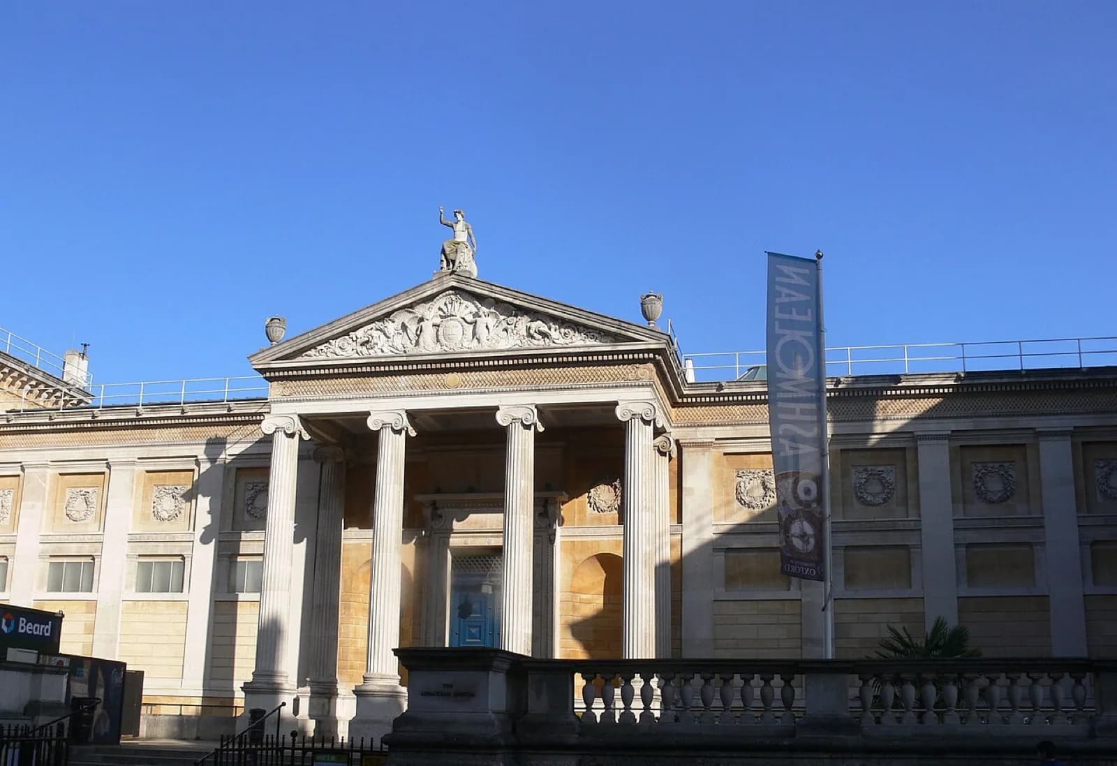 The grand neoclassical stone facade of the Ashmolean Museum on Beaumont Street in Oxford, United Kingdom, with ionic columns, triangular pediment bearing the name, flags flying, and people walking on the sidewalk beneath a clear blue sky.