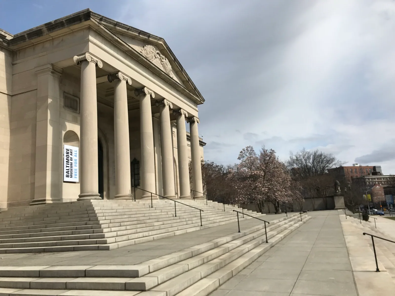 Neoclassical portico of the Baltimore Museum of Art designed by John Russell Pope, featuring tall fluted columns, a sculpted pediment, broad stone staircase with 'Free for All' banner, trees, and surrounding plaza.