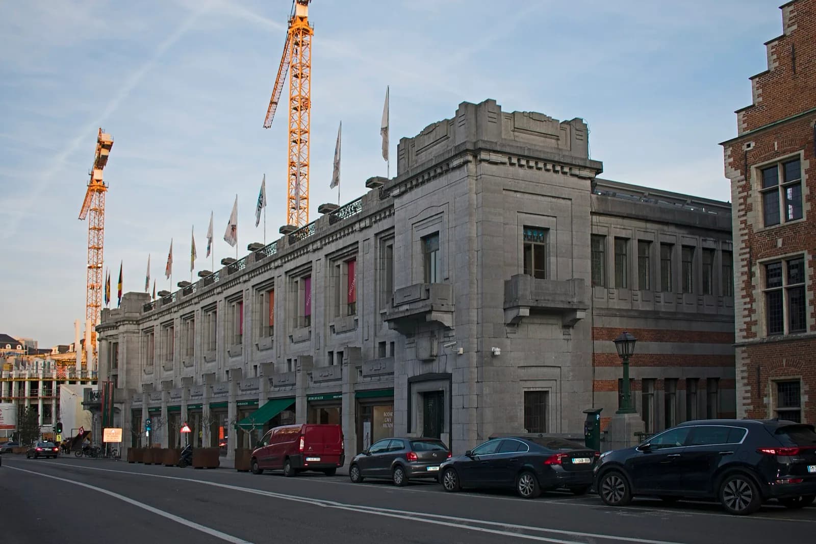 The main stone facade of BOZAR (Palais des Beaux-Arts), an Art Deco building by Victor Horta on Rue Ravenstein in Brussels, featuring symmetrical architecture, large windows, decorative cornices, vertical flags and banners, with parked cars along the street and construction cranes visible in the background under a clear sky.