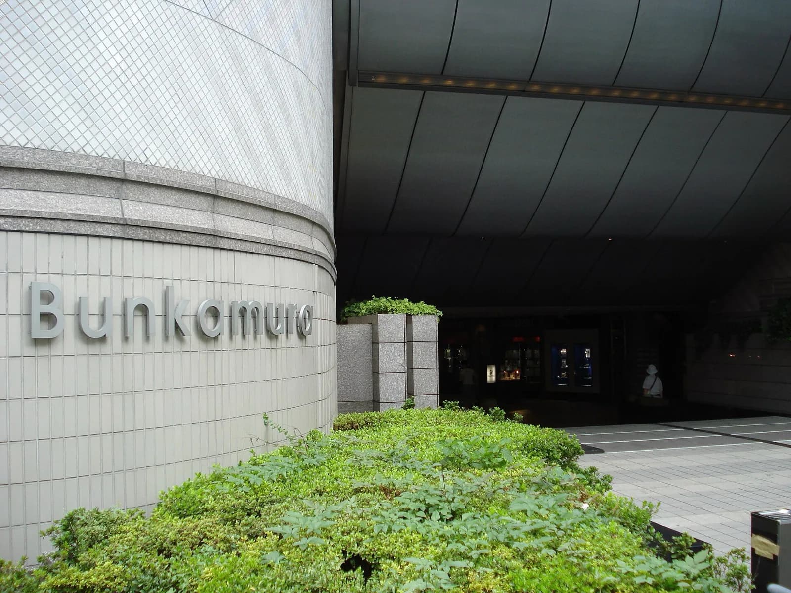 The curved modern facade of Bunkamura cultural complex in Shibuya, Tokyo, featuring large metallic lettering spelling 'Bunkamura', light gray cladding, and landscaped greenery at the entrance under an overhanging structure.