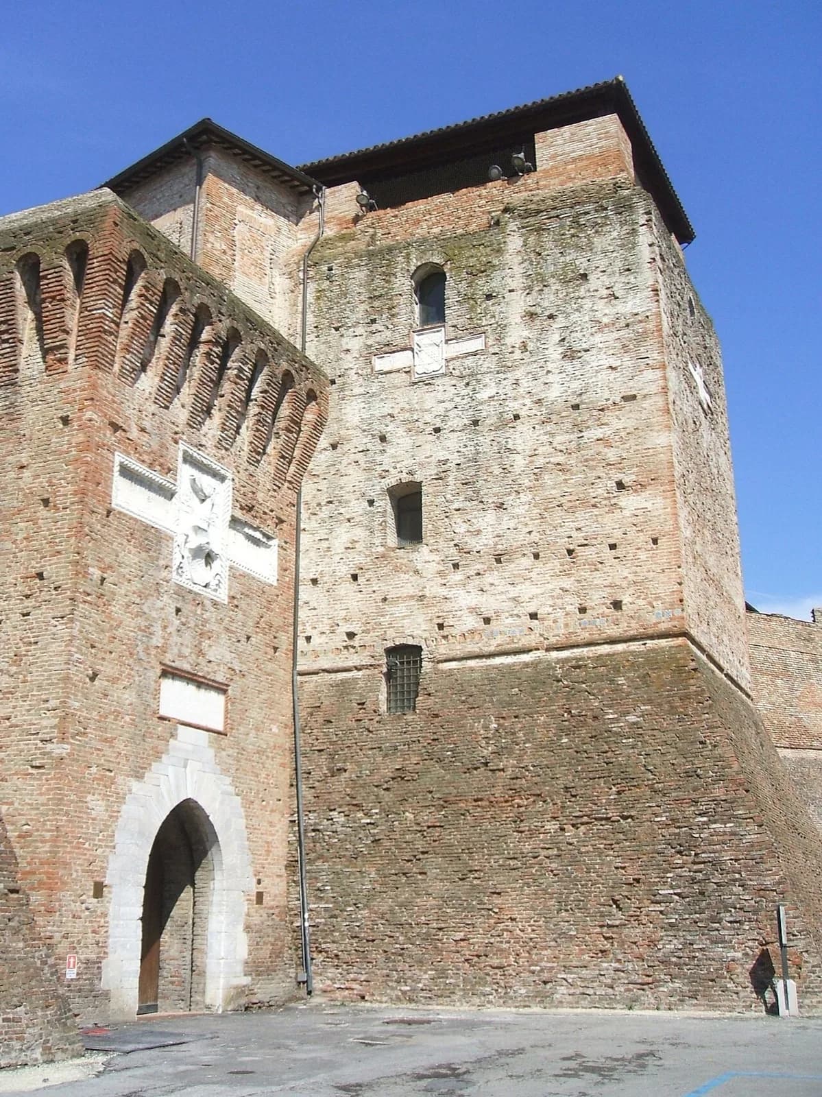 The imposing brick fortress of Castel Sismondo in Rimini, showing a tall weathered tower with brick battlements, stone relief coats of arms featuring eagles, narrow windows, and a large arched gateway entrance under a clear blue sky.