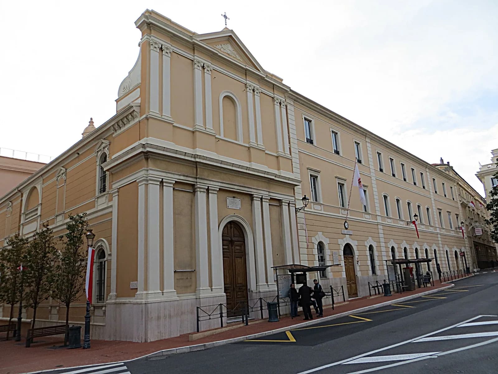 Corner view of the Musée de la Chapelle de la Visitation (Chapelle de la Visitation) in Monaco-Ville: a Baroque-style building with pale yellow stucco walls, white stone pilasters and moldings, an arched wooden entrance door, and a triangular pediment topped by a cross, located at a street intersection with pedestrians, Monaco flags, and a bus shelter.