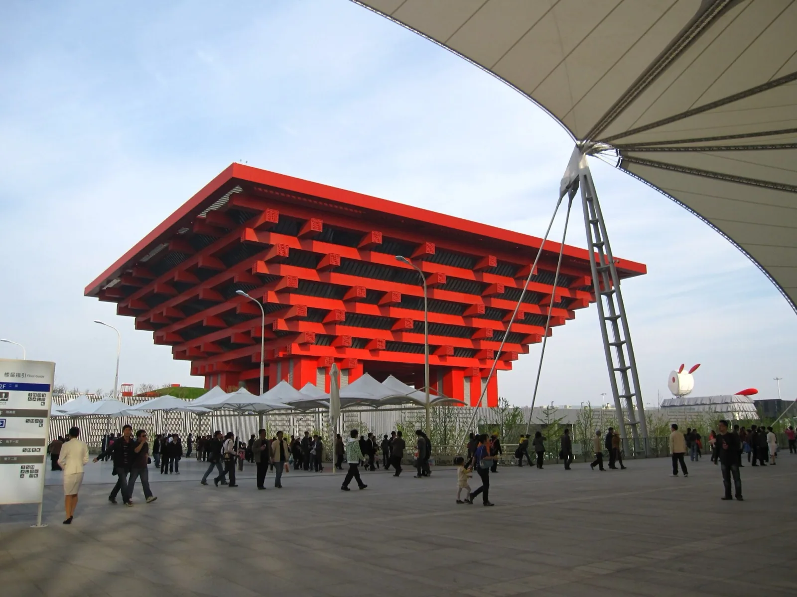 The iconic red inverted-pyramid China Pavilion (now China Art Museum / China Art Palace) at the former Expo 2010 site in Shanghai, a massive tiered structure with traditional Chinese architectural motifs, viewed across a crowded plaza with white shade structures under a clear sky.