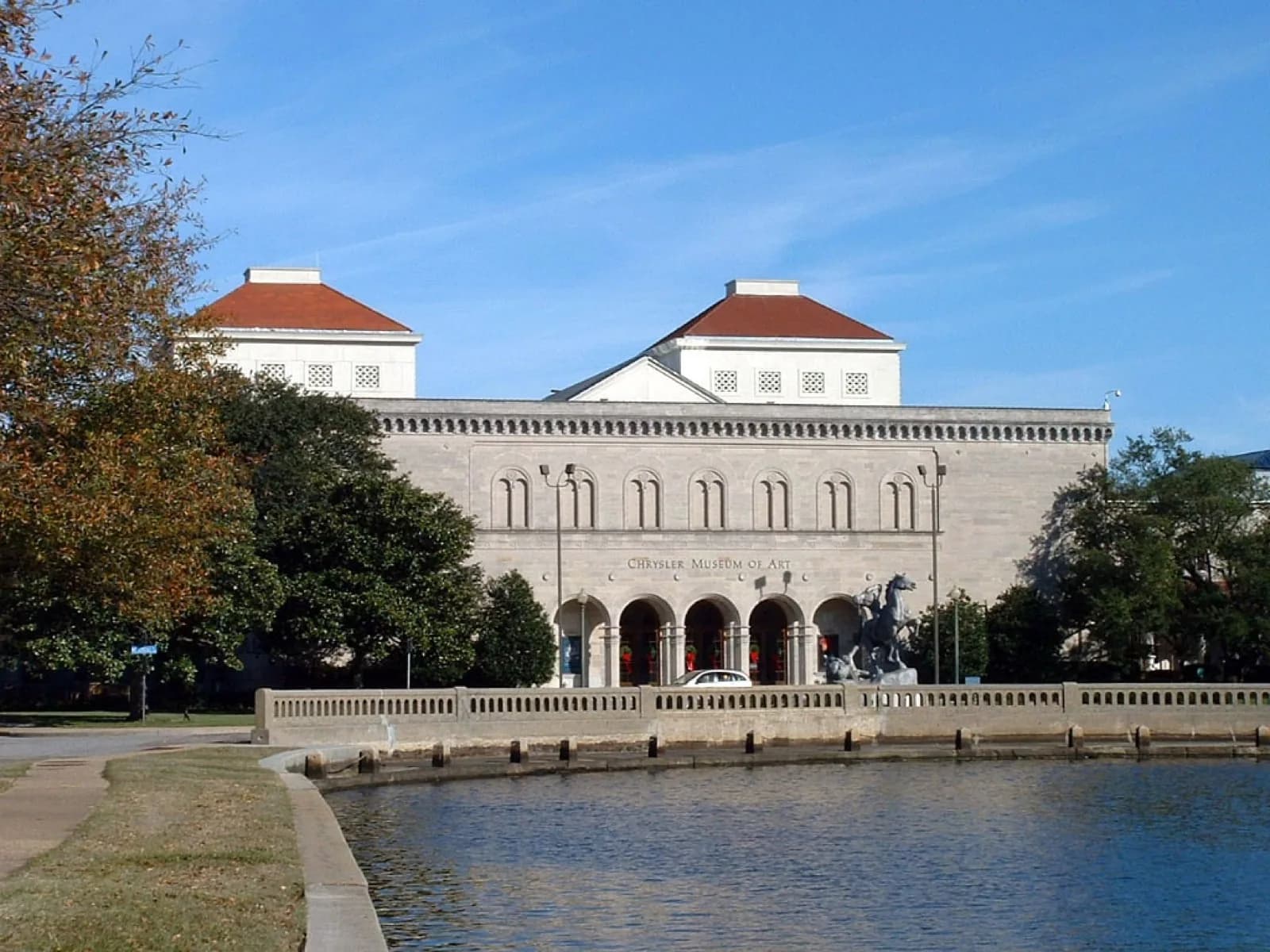 View across water of the Chrysler Museum of Art in Norfolk, Virginia, showing the grand stone building with red roofs, arched entrance, and equestrian sculpture under a clear blue sky.