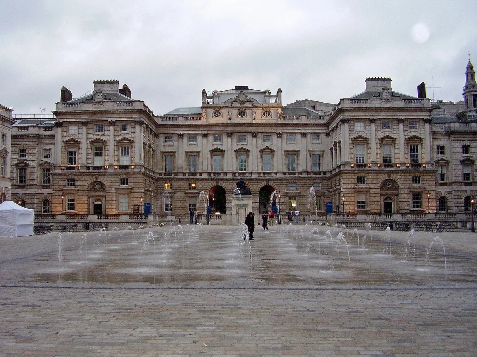 The neoclassical Strand block facade of Somerset House in London, home of the Courtauld Gallery, viewed across the Edmond J. Safra Fountain Court with its fountains and wet cobblestone paving under an overcast sky.
