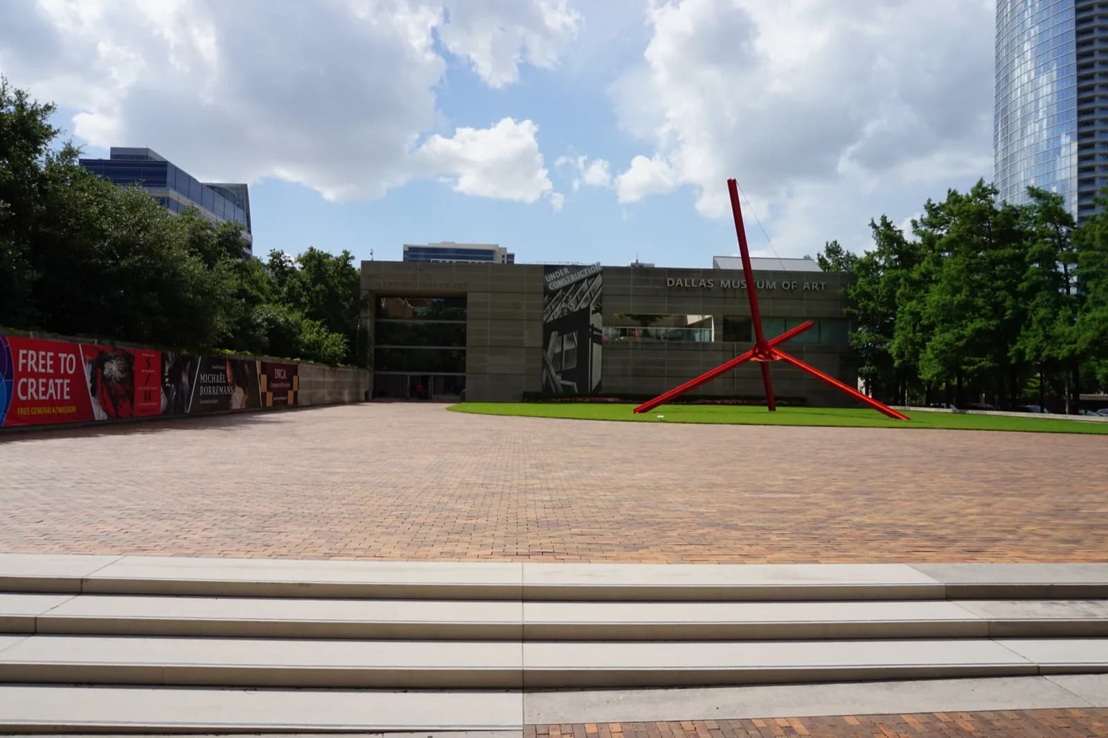 The modern glass and concrete facade of the Dallas Museum of Art, featuring large windows, geometric architecture, and a prominent entrance with public art in the foreground under a blue sky.