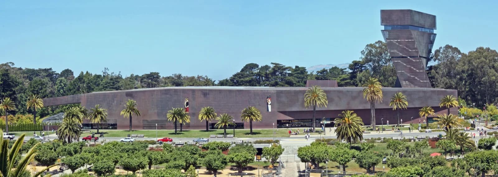Wide panoramic view of the de Young Museum in Golden Gate Park, San Francisco, featuring the distinctive copper-clad facade, angular observation tower, palm trees, and visitors under a clear blue sky.