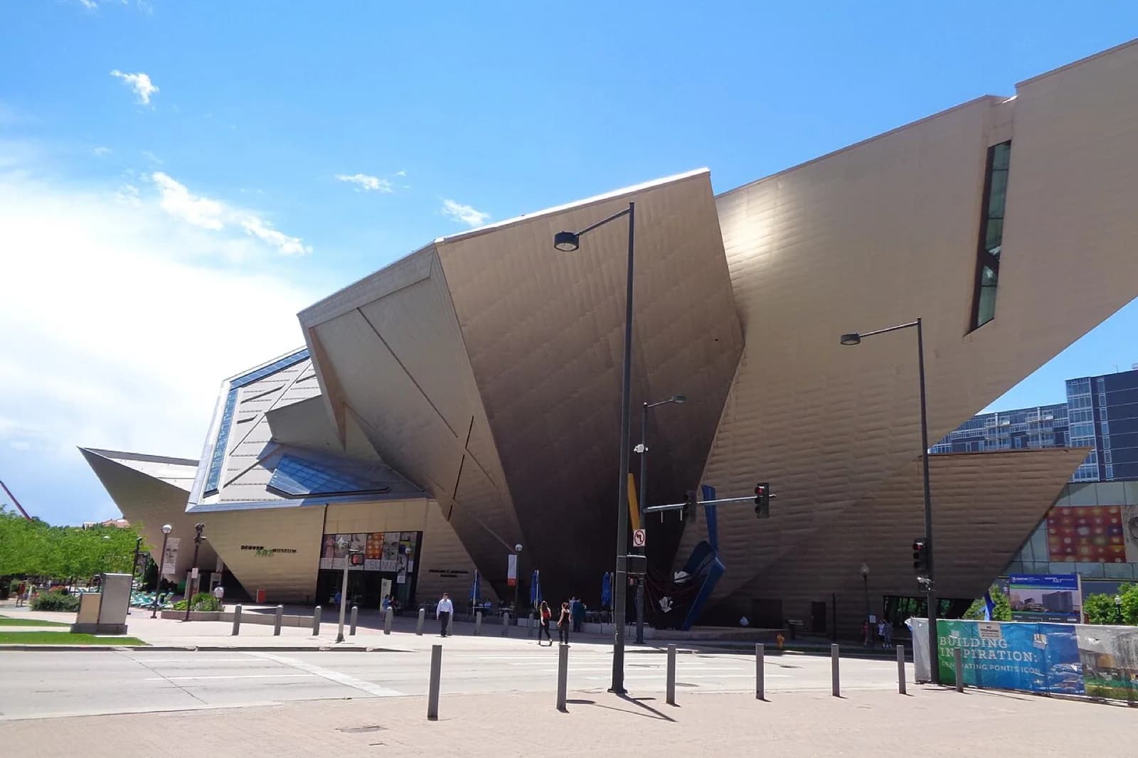 The Frederic C. Hamilton Building of the Denver Art Museum, a striking deconstructivist structure of angular titanium-clad forms with sharp peaks and geometric facets, viewed across the entrance plaza with steps, visitors, and flags on a clear day.