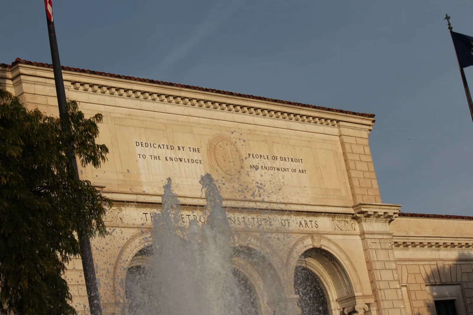 Grand stone facade of the Detroit Institute of Arts with prominent central inscription 'Dedicated by the People of Detroit to the Knowledge and Enjoyment of Art', large arched entrance, splashing water fountain in the foreground, flags, and trees under a clear blue sky.