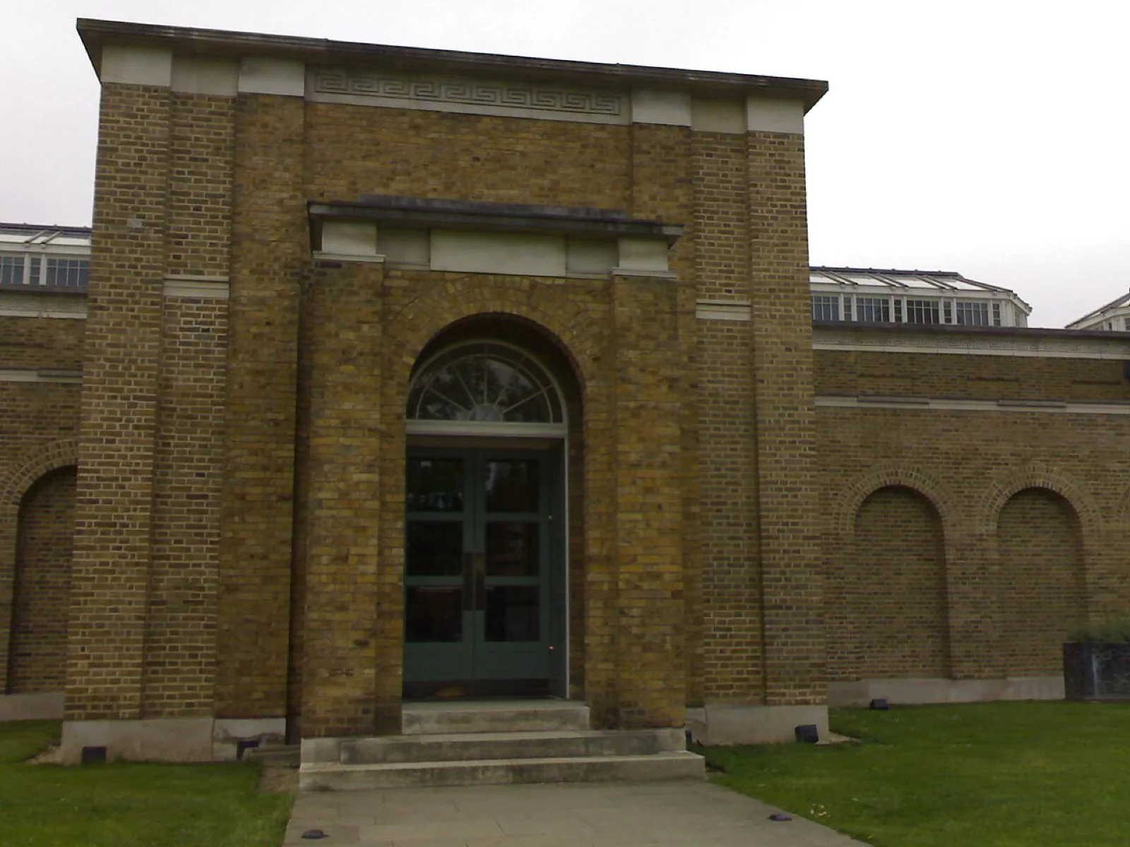 The neoclassical yellow-brick facade of the Dulwich Picture Gallery in London, designed by Sir John Soane, featuring a grand portico with columns, a pediment inscribed 'DULWICH PICTURE GALLERY', and a central dome, set against a blue sky with trees and visitors on the lawn.