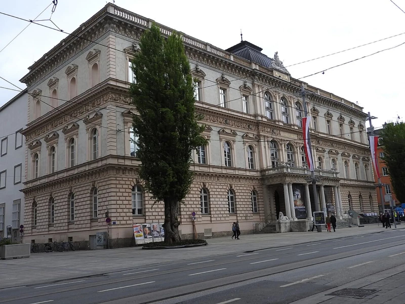 The ornate historic facade of the Tiroler Landesmuseum Ferdinandeum in Innsbruck, Austria, a grand Beaux-Arts style building with columns, decorative stonework, and entrance steps, viewed from the street with tram tracks and pedestrians.
