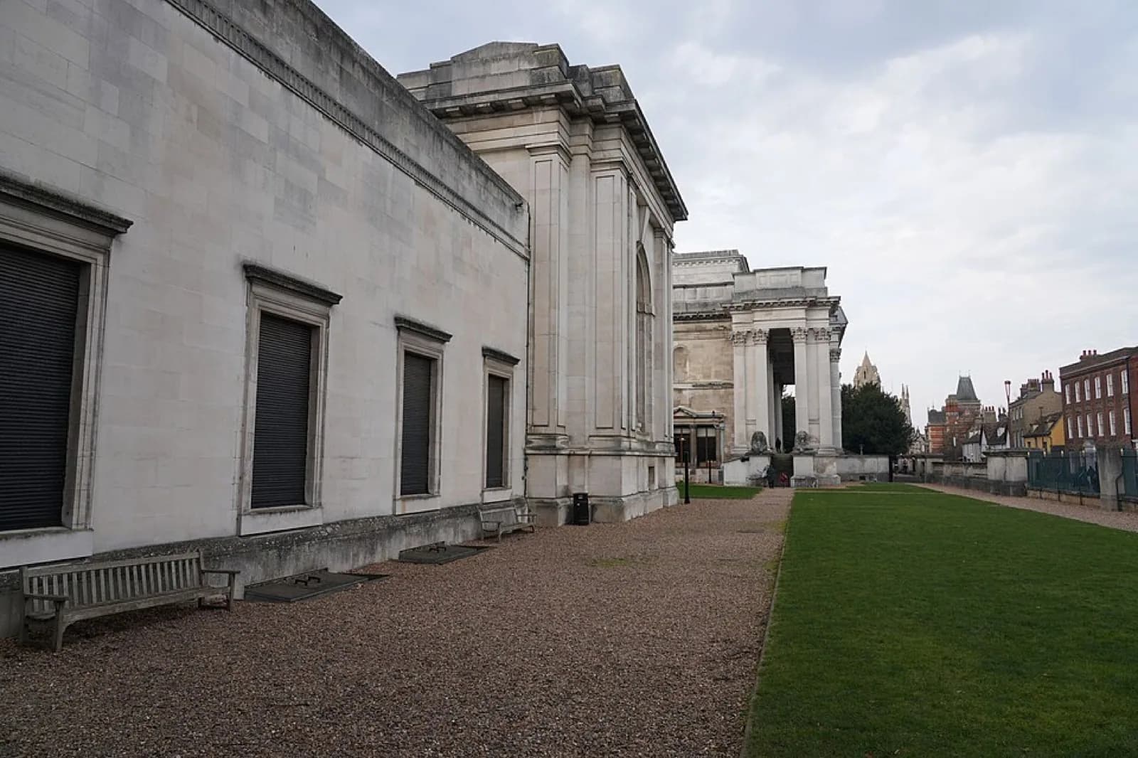 The side facade of the Fitzwilliam Museum in Cambridge, a neoclassical stone building with tall columns and pediment, viewed along a gravel path beside a lawn with benches under an overcast sky.