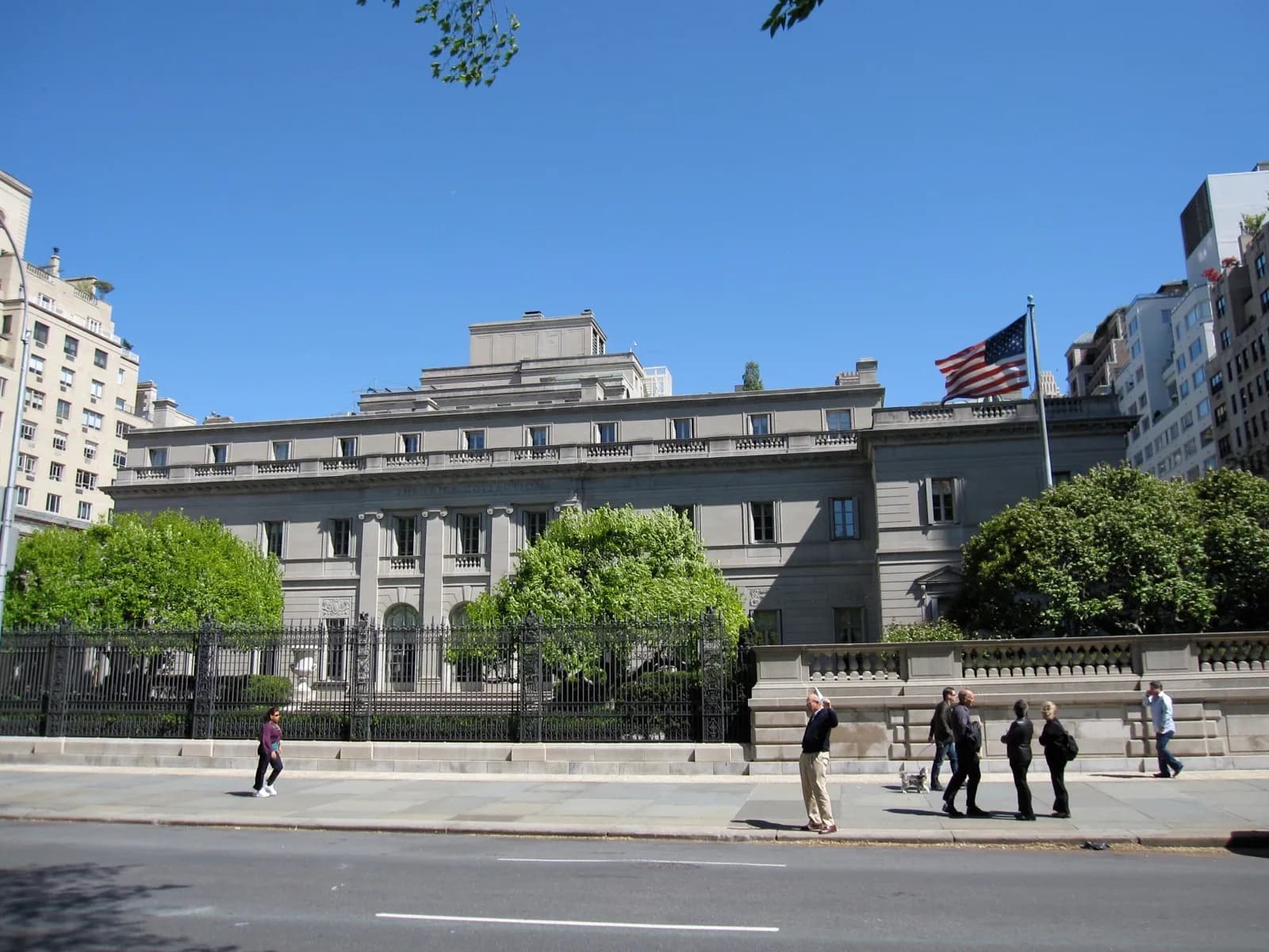 The Henry Clay Frick House on Fifth Avenue in New York City, home of the Frick Collection, a grand limestone Beaux-Arts mansion with classical architecture and an American flag, viewed from the street on a sunny day.