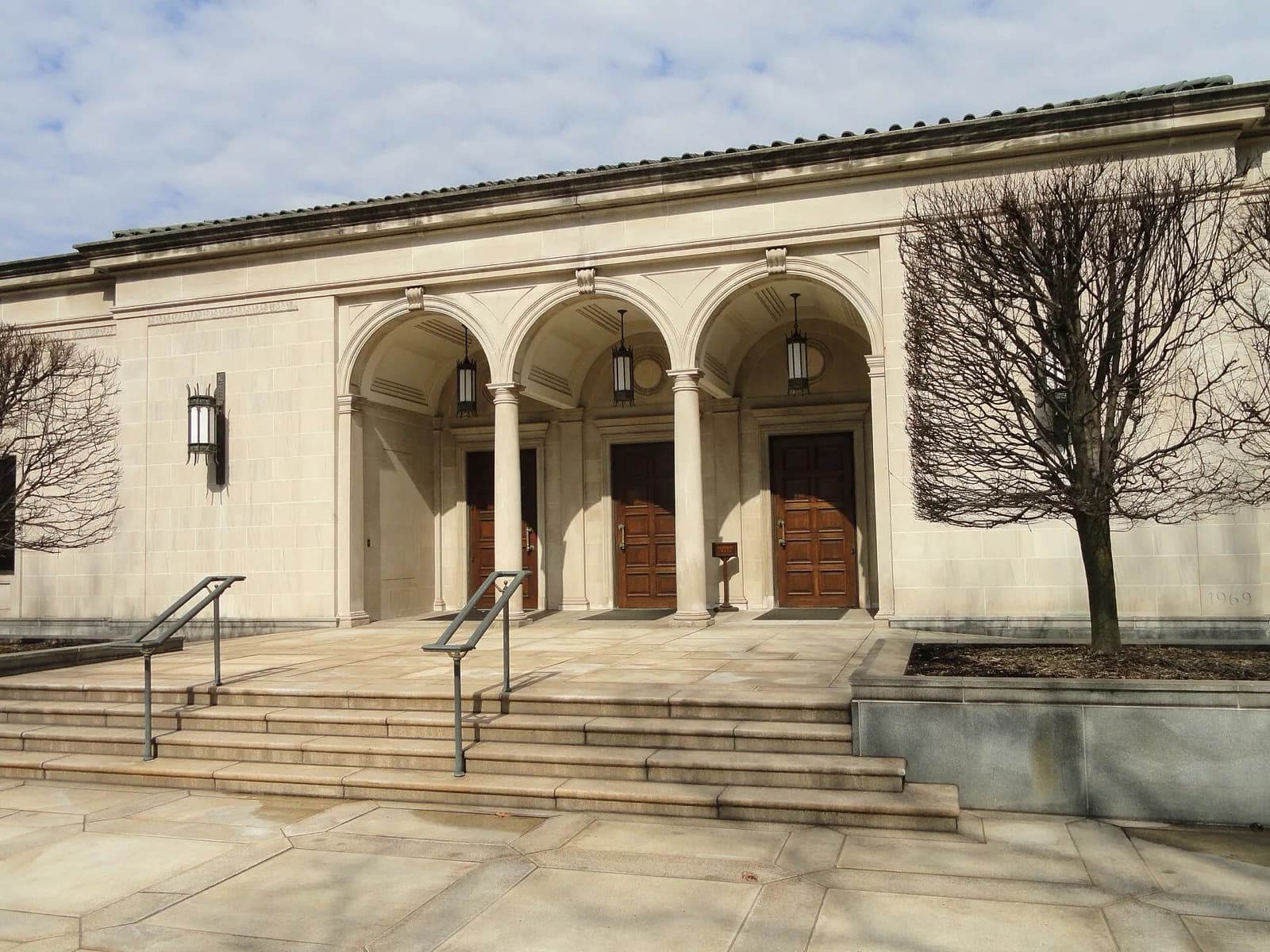 The Frick Art Museum at The Frick Pittsburgh, a classical limestone building with a grand arched portico and columns, wooden entrance doors, lanterns and steps, set on the former Frick estate with bare trees in the foreground under a partly cloudy sky.