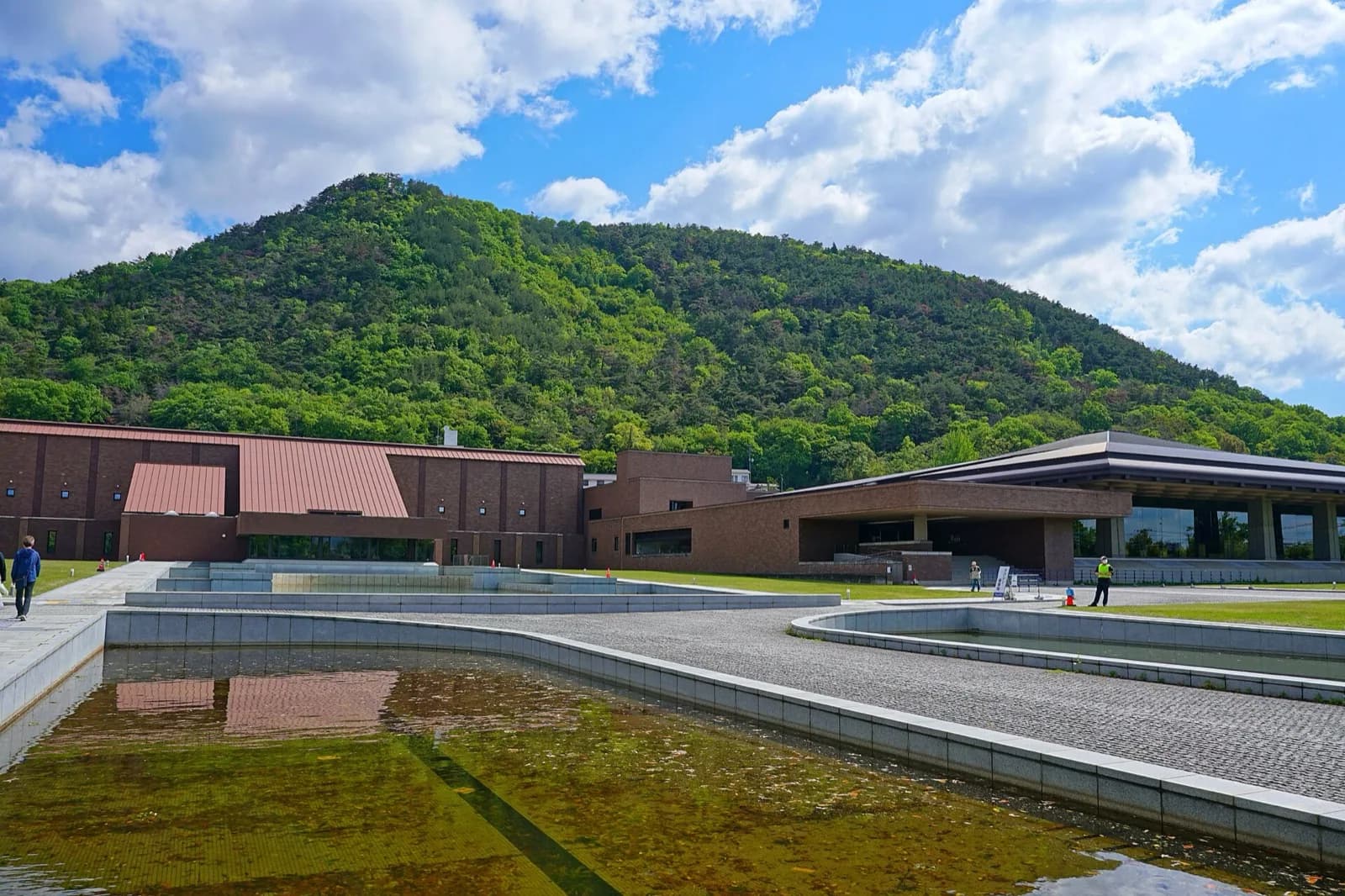 The modern brick and red-roofed buildings of the Fukushima Prefectural Museum of Art complex in Japan, with reflecting pools in the foreground and the green forested Mount Shinobu rising behind under a blue sky with white clouds.