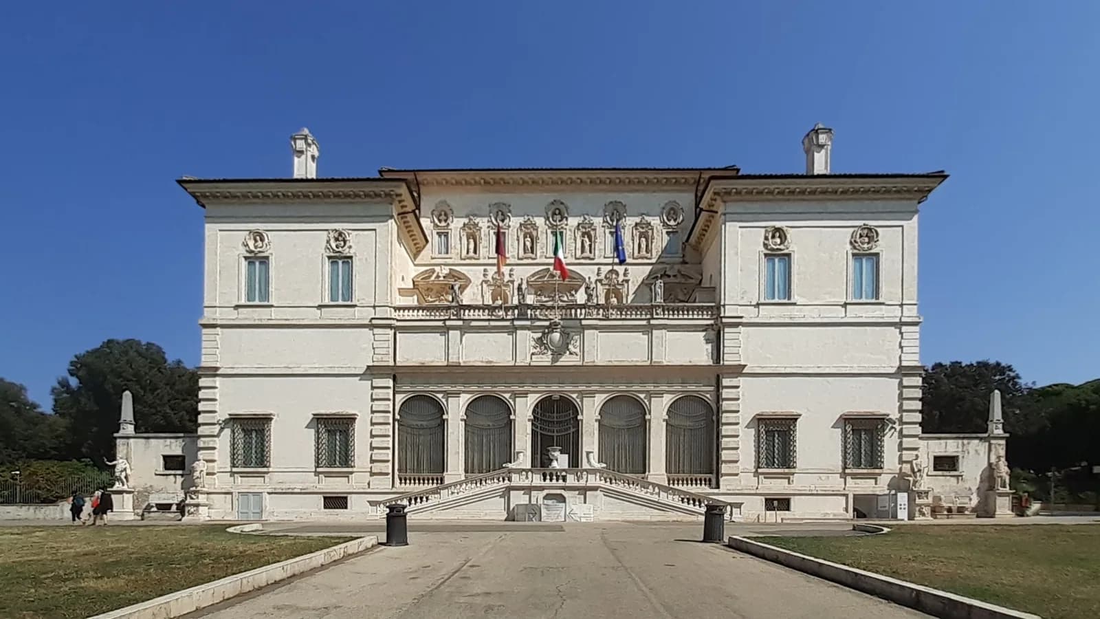 Symmetrical facade of the Villa Borghese Pinciana housing the Galleria Borghese in Rome, a grand white Baroque-style palace with central staircase, arched ground floor, ornate upper level with niches and busts, Italian and EU flags, and surrounding park grounds under clear blue sky.