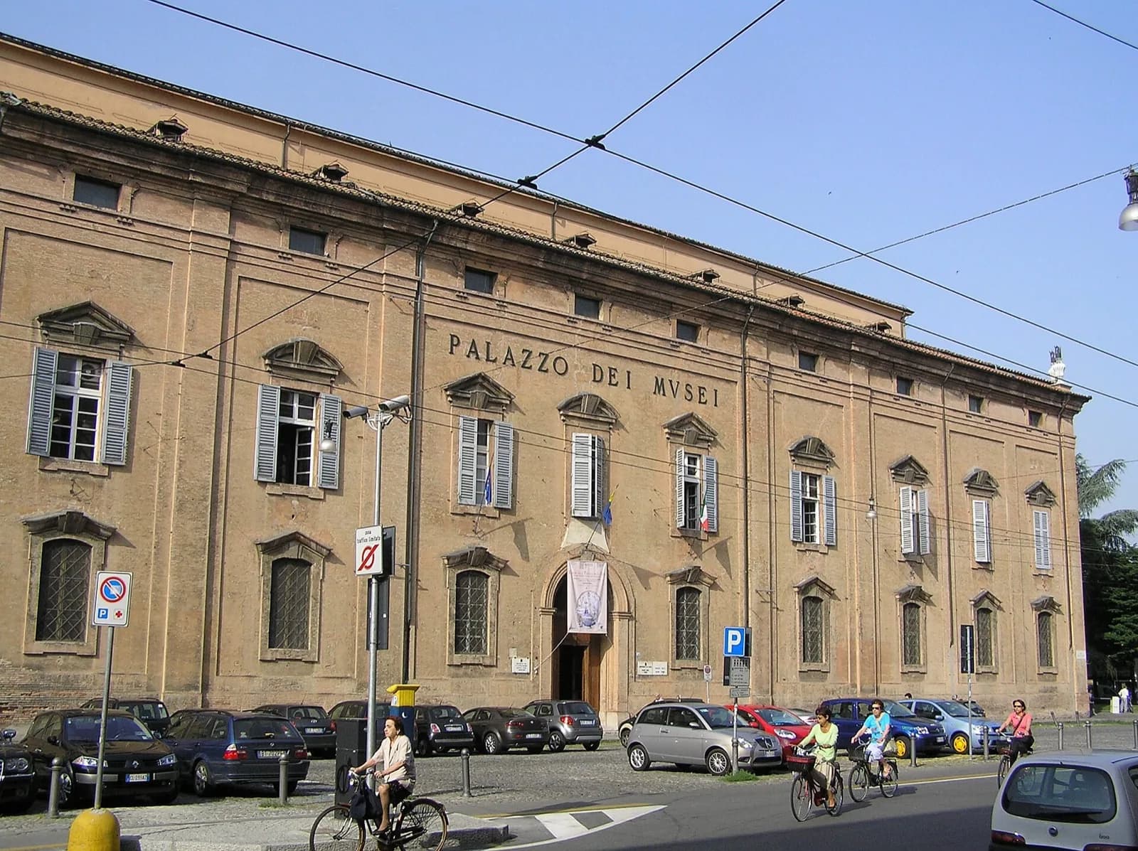 Facade of the Palazzo dei Musei in Modena, Italy, housing the Galleria Estense, seen from the street: a large historic ochre building with arched windows, shutters, central entrance and Italian flags, with cyclists, parked cars and blue sky in foreground.