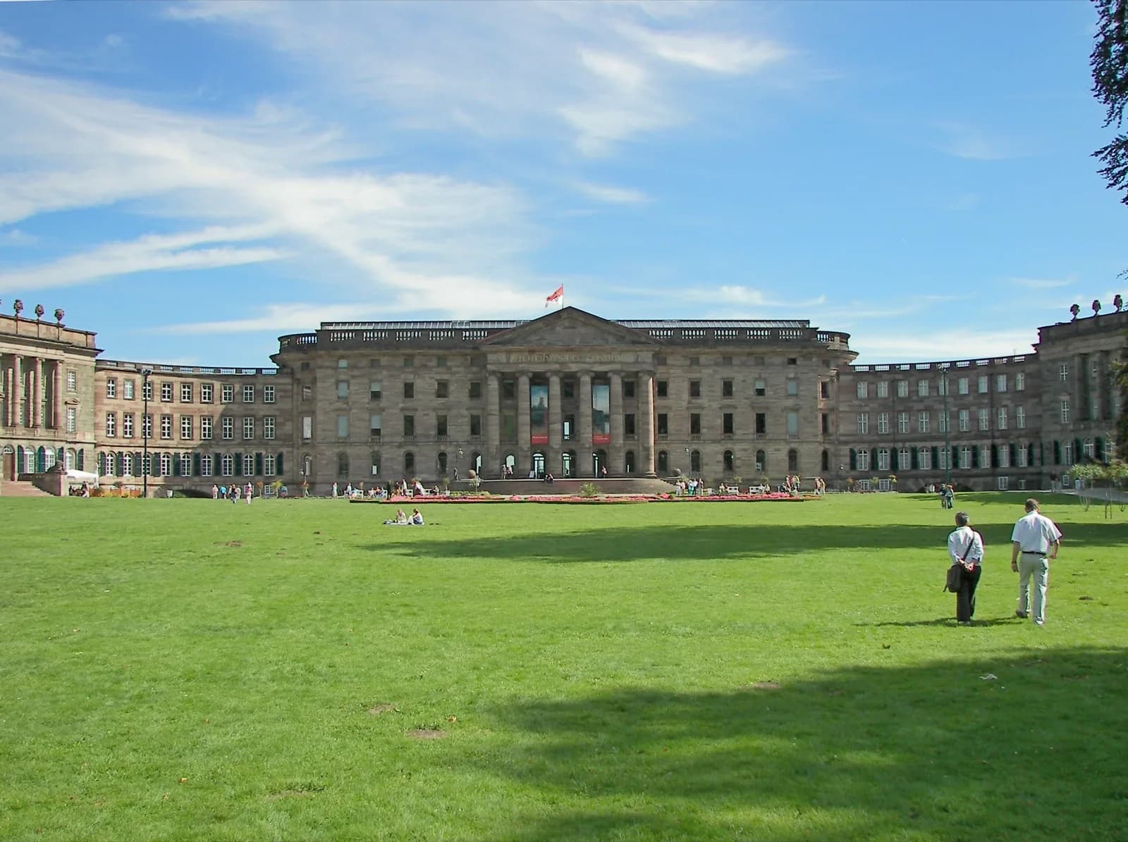 The Schloss Wilhelmshöhe in Kassel, Germany, a grand neoclassical palace with central columned portico and long wings housing the Gemäldegalerie Alte Meister, viewed across the expansive lawn with visitors under a blue sky.