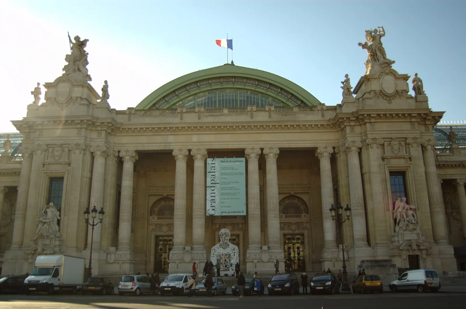The ornate Beaux-Arts stone facade of the Grand Palais in Paris, featuring grand columns, pediments, allegorical sculptures, and a large arched entrance with glass dome above, on the Champs-Élysées.