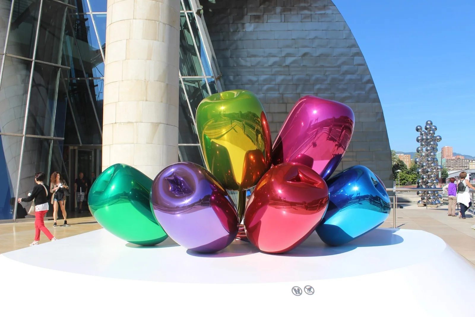 The titanium-clad, organically curved facade of the Guggenheim Museum Bilbao designed by Frank Gehry, with a large rainbow-colored metal tulip sculpture by Jeff Koons in the foreground under a bright blue sky.