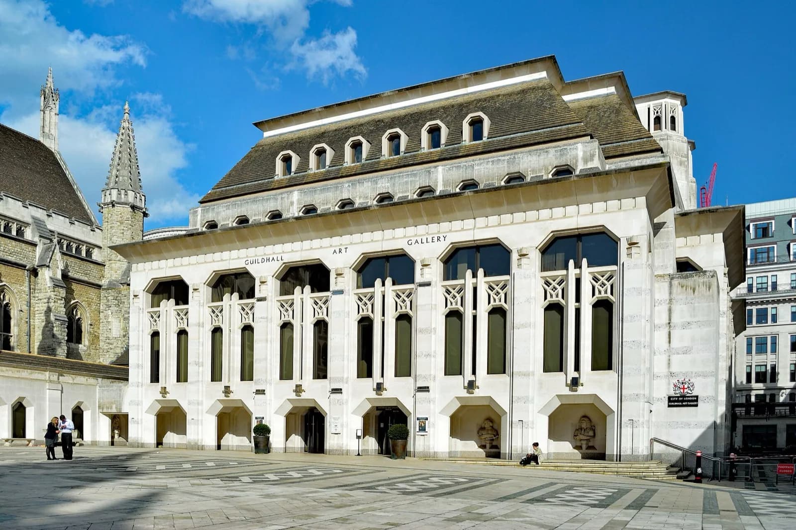 The white stone facade of the Guildhall Art Gallery in London, featuring tall arched windows with geometric grilles, inscribed 'GUILDHALL ART GALLERY', and sculpted figures at the base, set in a paved plaza beside the historic Guildhall tower and spire under a bright blue sky.