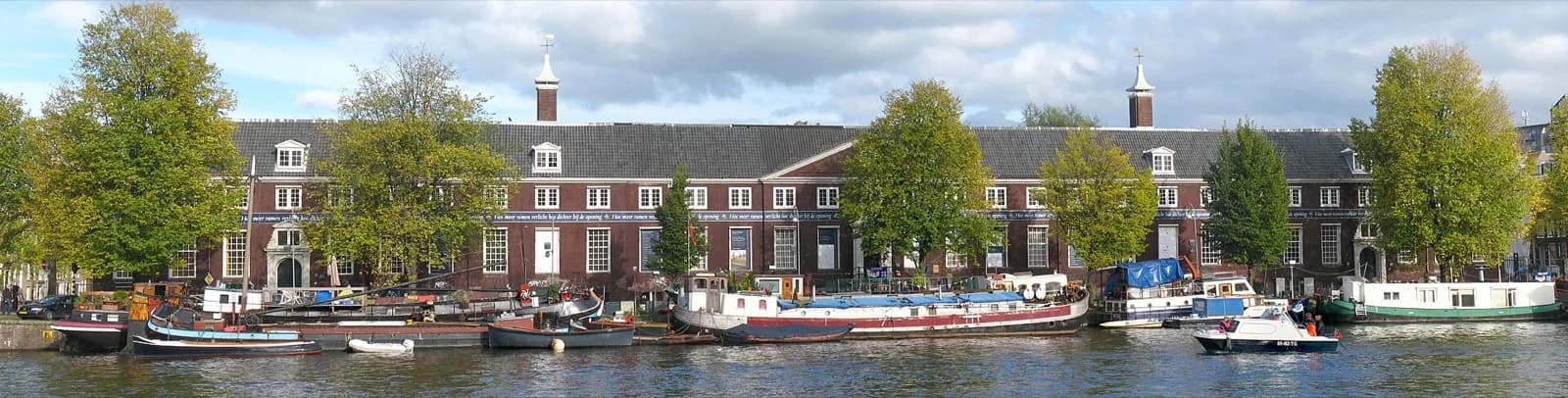 The long classical red-brick facade of the former Amstelhof, now H'ART Museum, along the Amstel river in Amsterdam, with its symmetrical windows and gables, viewed across the water with boats and trees on a cloudy day.