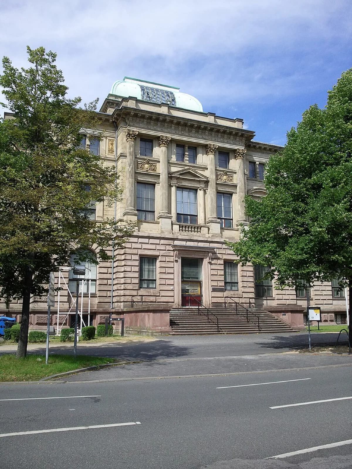 The classical stone facade of the Herzog Anton Ulrich-Museum in Braunschweig, Germany, a grand historic building with tall columns, ornate pediment, arched windows, and a central staircase entrance, set against trees and a blue sky.