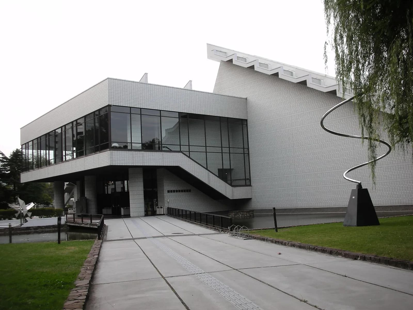 The modern white-tiled exterior of the Hokkaido Museum of Modern Art in Sapporo, Japan, featuring large glass windows on the upper level, an external staircase, a paved plaza, abstract metal sculptures including a prominent spiral, grass lawns, and a water feature under a bright sky.