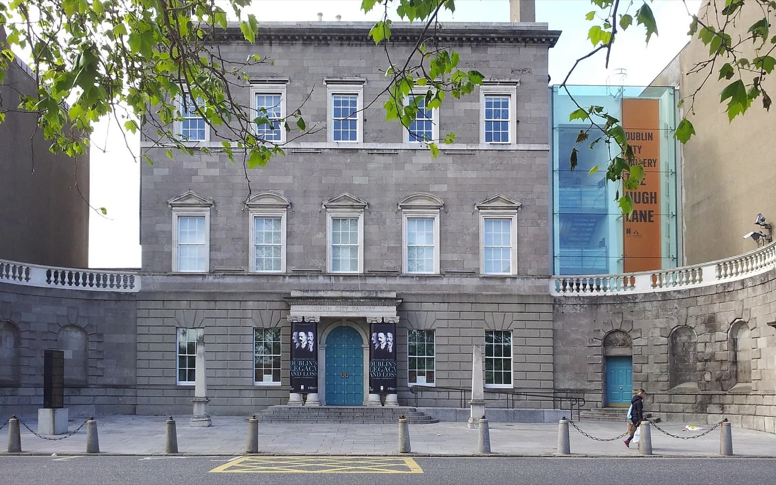 The neoclassical gray stone facade of Charlemont House, home to the Hugh Lane Gallery in Dublin, featuring a central teal door under a portico, symmetrical sash windows, banners for a Francis Bacon exhibition, and a modern glass extension with the gallery's orange signage on the right, viewed from Parnell Square on a bright day with trees and a pedestrian in the foreground.