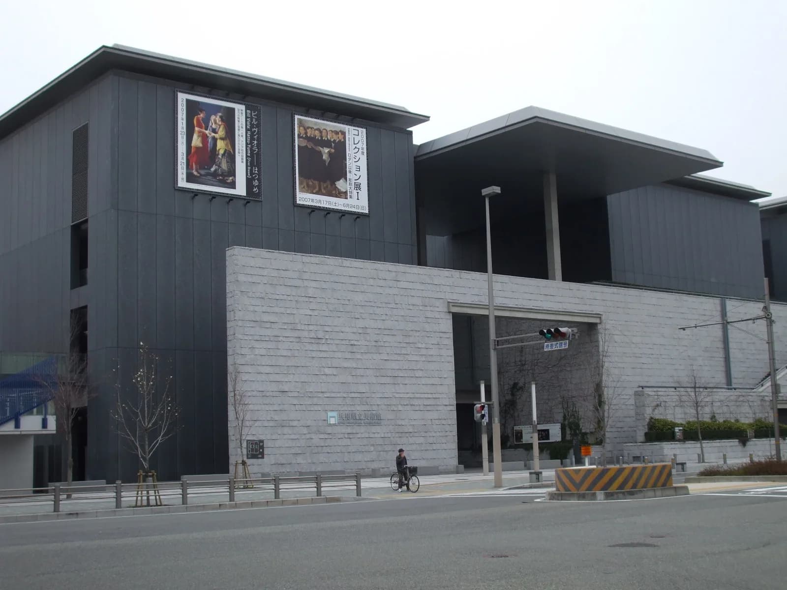 The Hyogo Prefectural Museum of Art in Kobe, Japan, a Tadao Ando-designed concrete structure with dark and light stone facades, large exhibition banners on the wall, an arched entrance, and a street-level view including a cyclist and traffic signals.