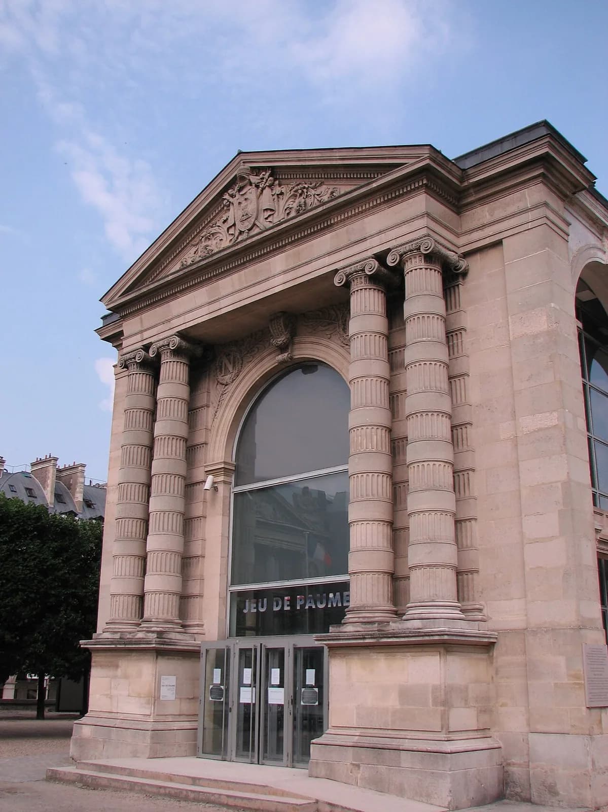 The classical stone facade of the Galerie Nationale du Jeu de Paume in the Tuileries Gardens, Paris, featuring tall columns, a triangular pediment, the inscription 'GALERIE NATIONALE DU JEU DE PAUME' above the entrance, surrounded by trees and visitors on a sunny day with blue sky.