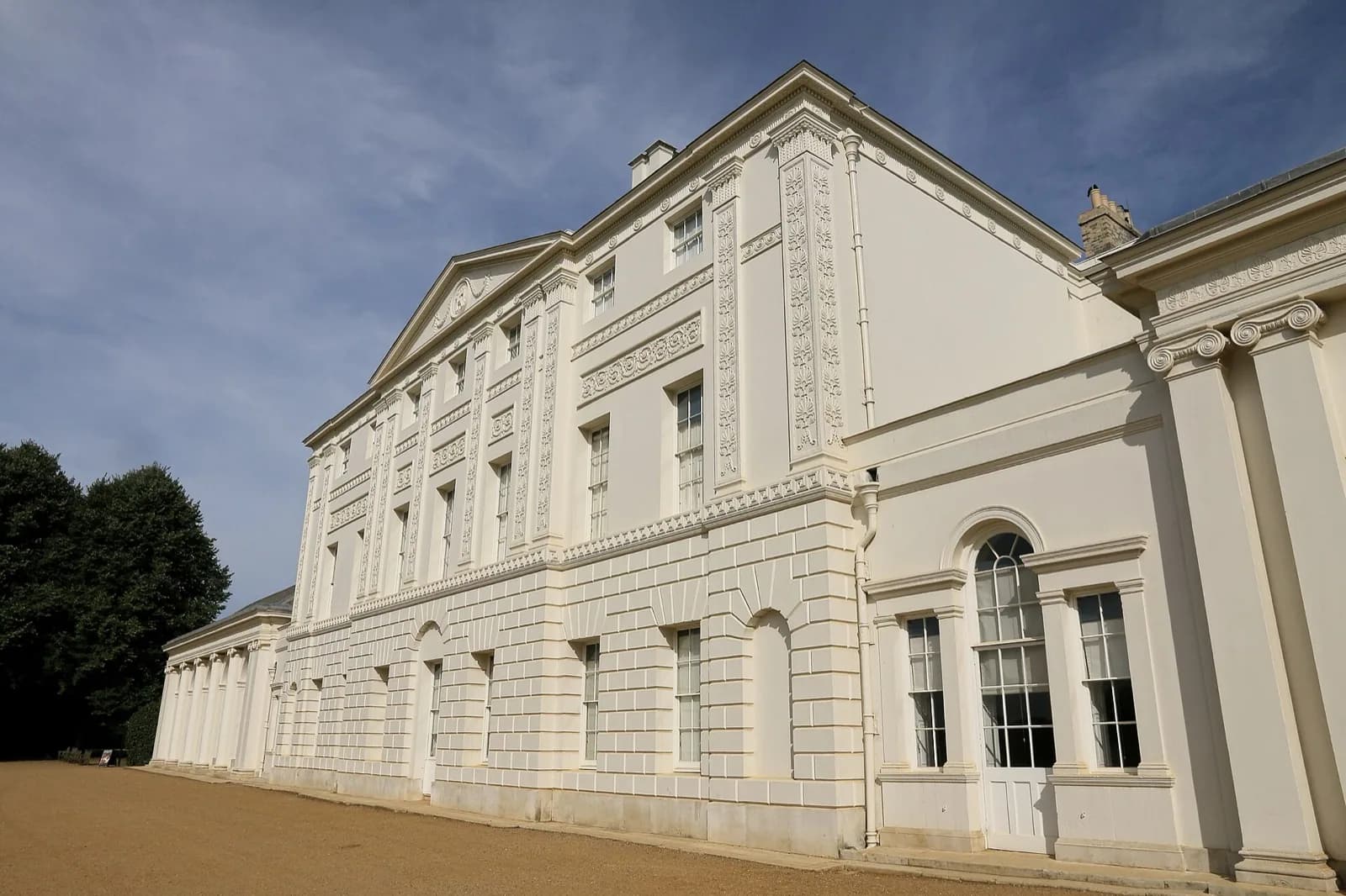 Kenwood House, a grand white neoclassical villa with a pedimented Ionic portico and symmetrical wings, set amid expansive green lawns and mature trees on the edge of Hampstead Heath in London under a bright blue sky; visitors visible on the grounds.