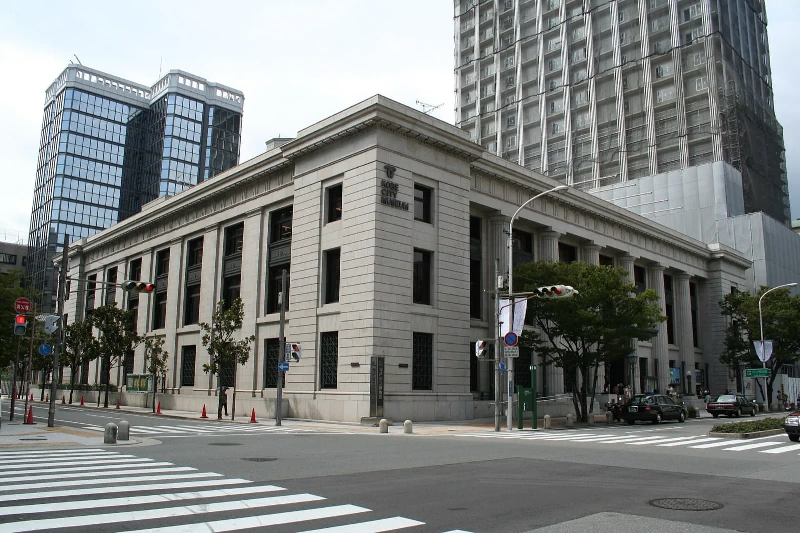 The Kobe City Museum, housed in a historic neoclassical stone building with tall gridded windows and columns at a street intersection in Kobe, Japan, with modern skyscrapers, traffic, and trees in the background under an overcast sky.