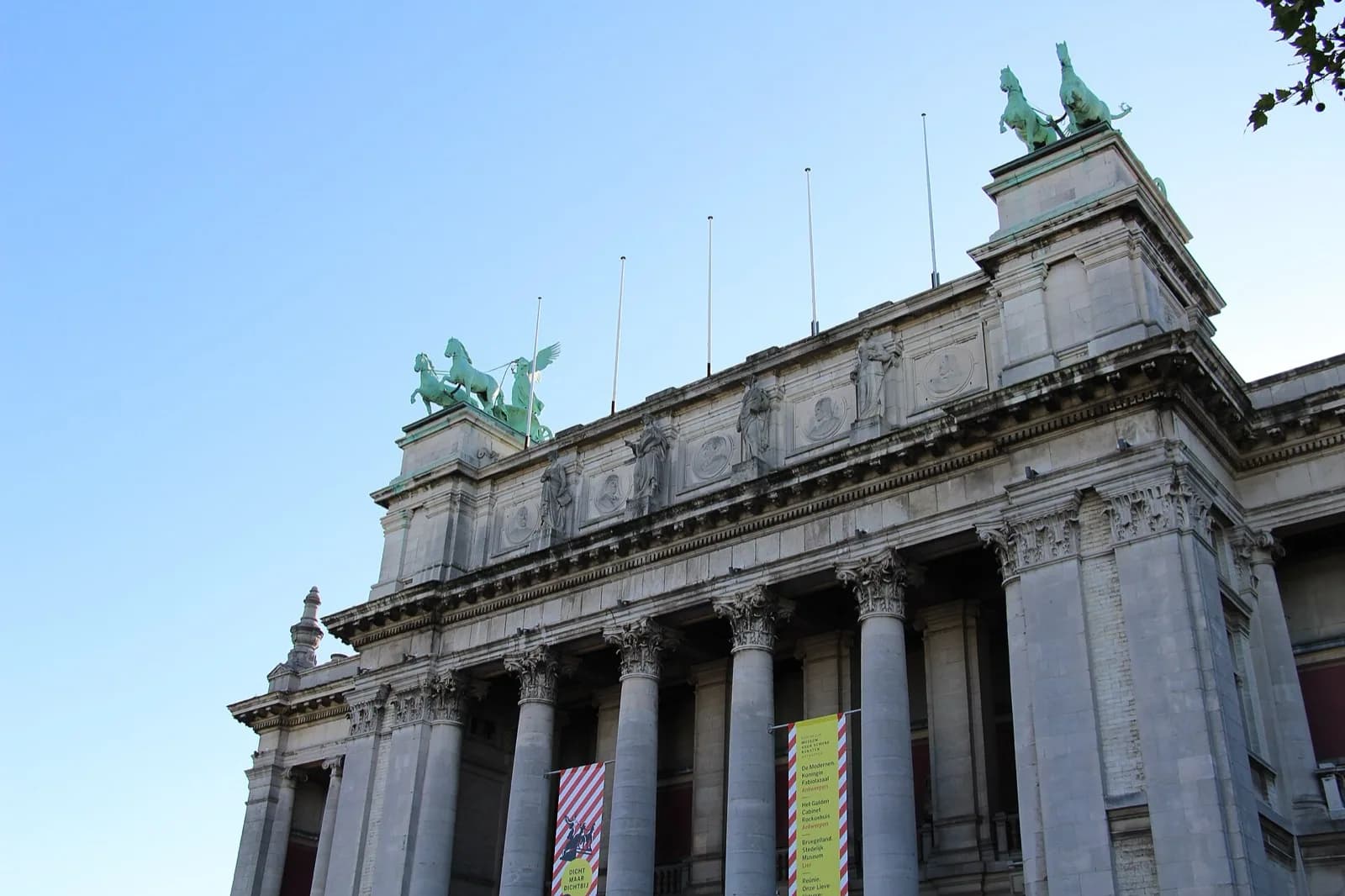 The grand neoclassical facade of the Koninklijk Museum voor Schone Kunsten (KMSKA) in Antwerp, Belgium, featuring a long row of tall columns, statues along the roof, a central dome, and a reflecting pool in front, under a clear blue sky with visitors.