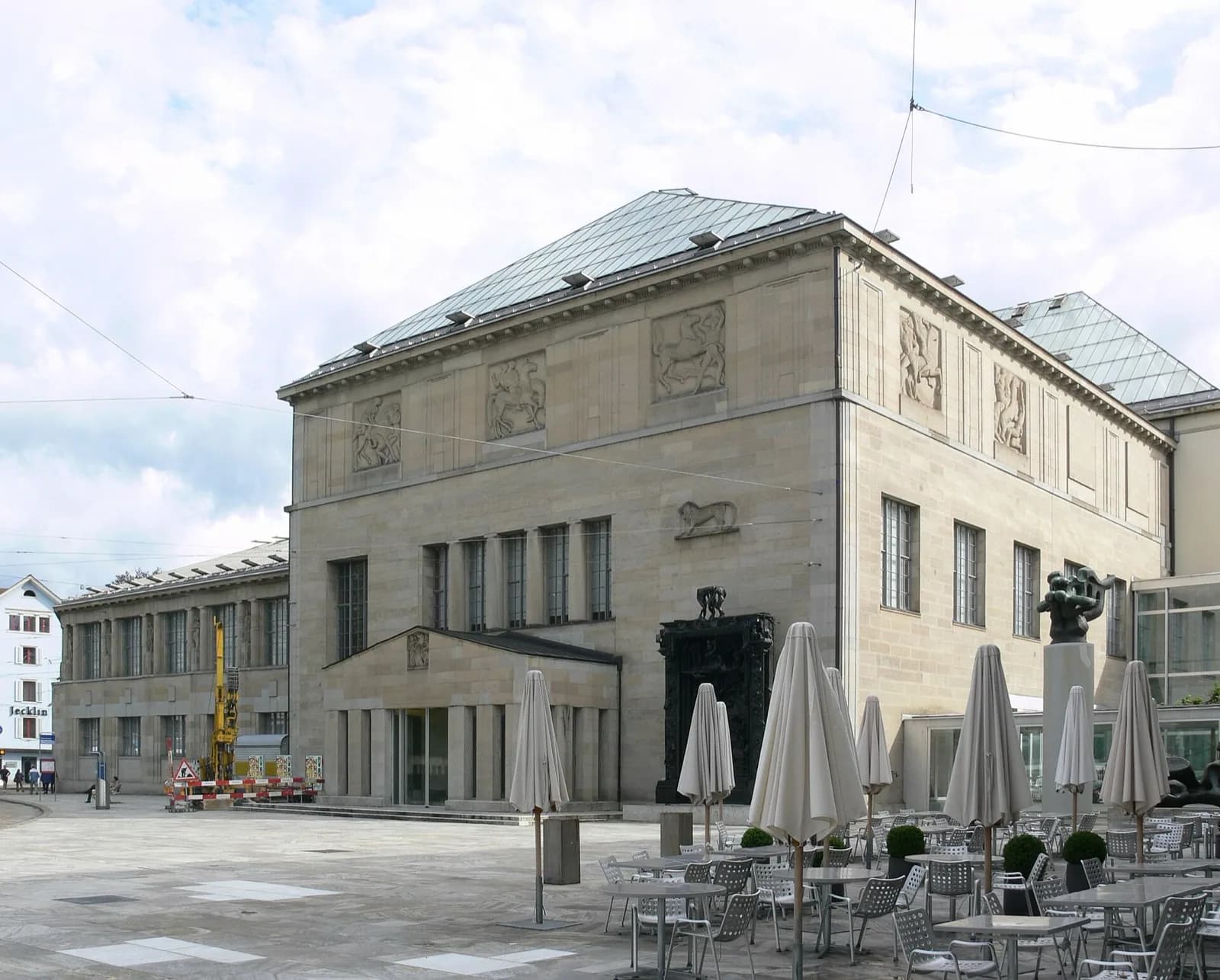 The light stone neoclassical facade of the Kunsthaus Zürich at Heimplatz, featuring relief sculptures, columns and pediment, with Auguste Rodin's monumental bronze 'La Porte de l'Enfer' at the entrance, outdoor cafe seating with umbrellas in the foreground plaza, under a bright partly cloudy sky in Zürich.