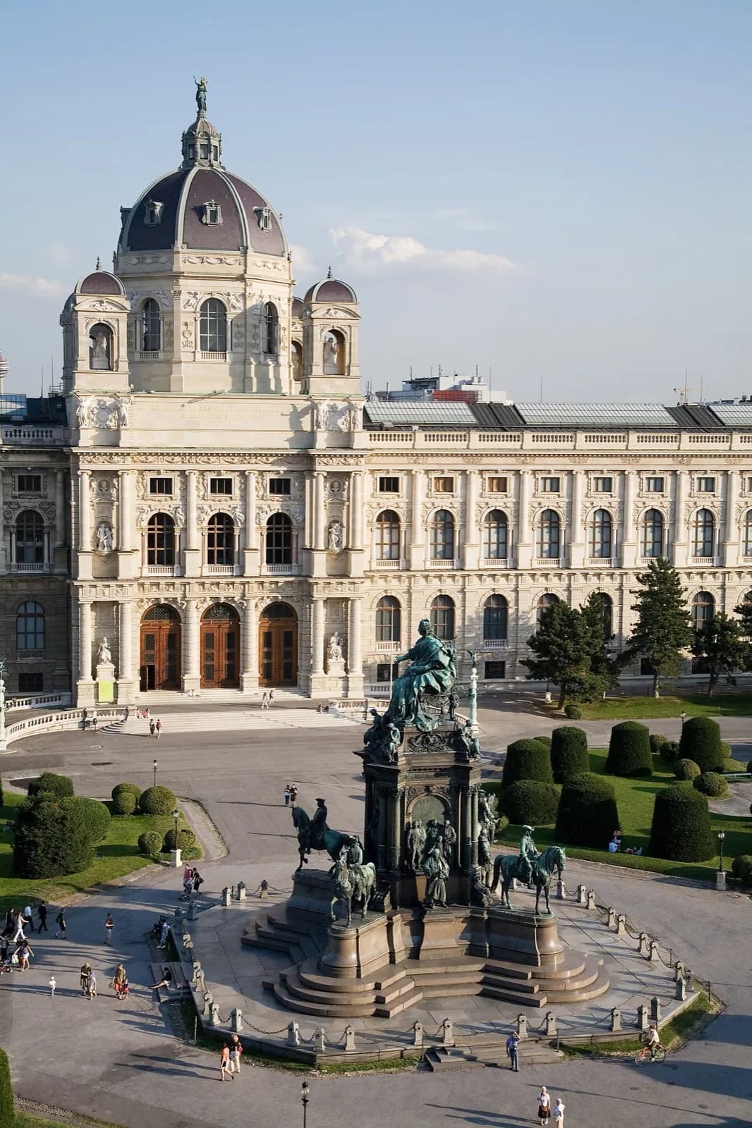 A high-angle view of the grand ornate facade of the Kunsthistorisches Museum in Vienna, a pale stone Renaissance Revival building with a prominent dome, arched windows and sculptures, facing Maria-Theresien-Platz with its central monumental equestrian sculpture, manicured topiary, and visitors on a sunny day.