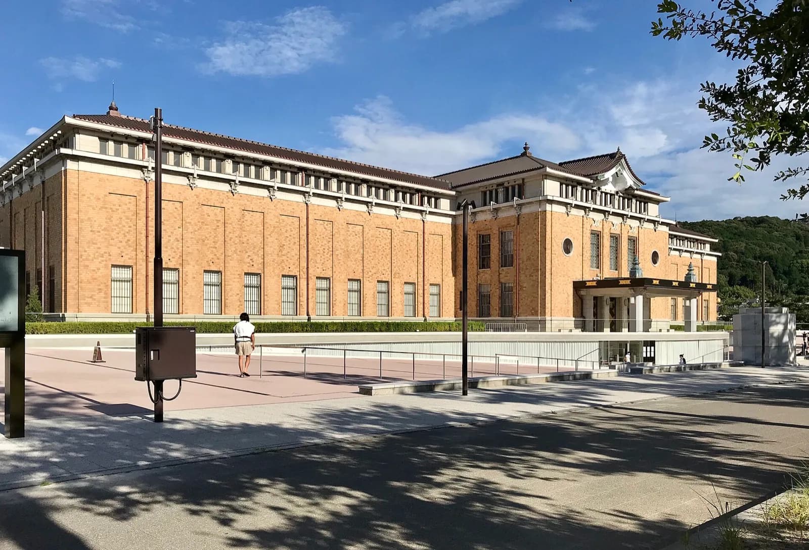 The Kyoto City KYOCERA Museum of Art (Kyoto Municipal Museum of Art) in Kyoto, Japan, a large brick building combining Western architecture with traditional Japanese tiled roofs and ornate details, viewed across the plaza under a blue sky with scattered clouds.