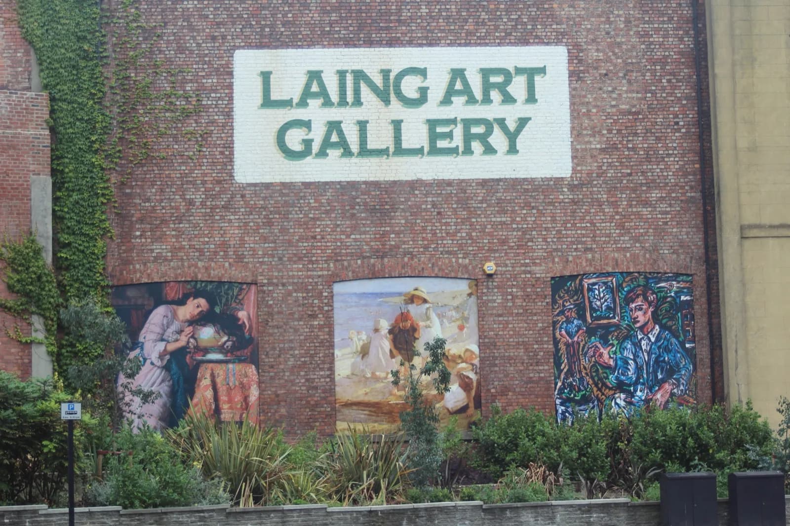 The red brick exterior of the Laing Art Gallery in Newcastle upon Tyne, featuring a prominent white 'LAING ART GALLERY' sign, three large arched mural paintings on the facade depicting historical and modern scenes, with greenery and the 'Blue Carpet' public space in the foreground.