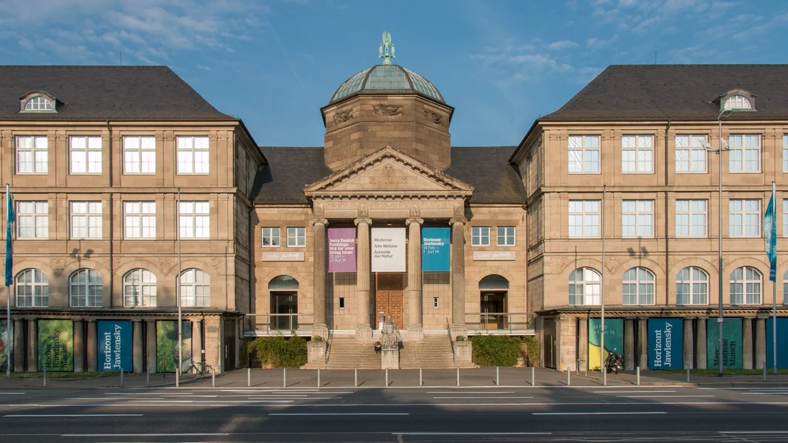 The grand neoclassical stone facade of the Museum Wiesbaden with its central columned portico, pediment, and copper dome under a clear blue sky, featuring exhibition banners and a statue in front.
